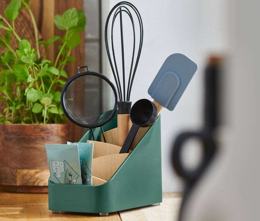 Ein grüner Utensilienständer beinhaltet ein Sieb, einen Schneebesen, einen Messbecher, einen Spatel und eine Packung Tee.
