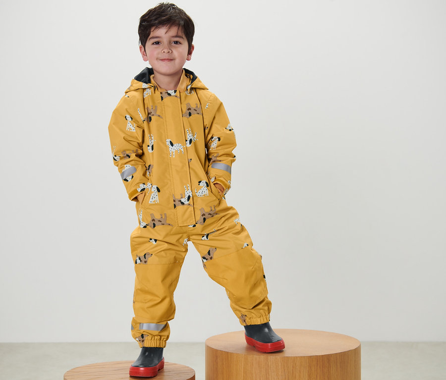 Ein Junge steht auf einem Holzsockel und trägt einen gelben Regen-Einteiler mit Hundeprint.