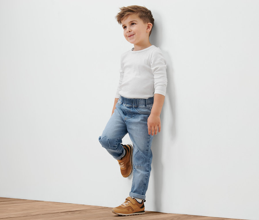 Ein Junge in einem weißen T-Shirt und Kleinkind-Schlupfdenim mit Häuschen-Stickerei lehnt an einer weißen Wand.