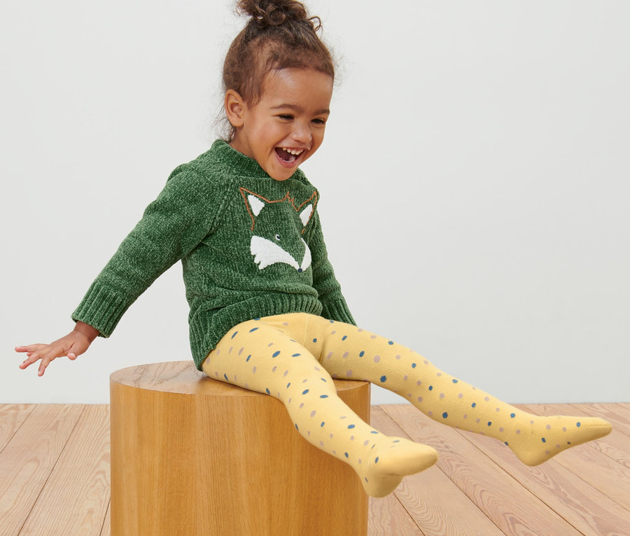 Mädchen sitzt auf einem Holzhocker und trägt 5 Kleinkind-Strumpfhosen.