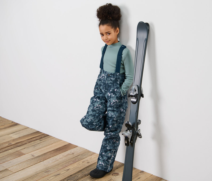 Mädchen in Kinder-Skihose mit Alloverprint, die an Ski lehnt.