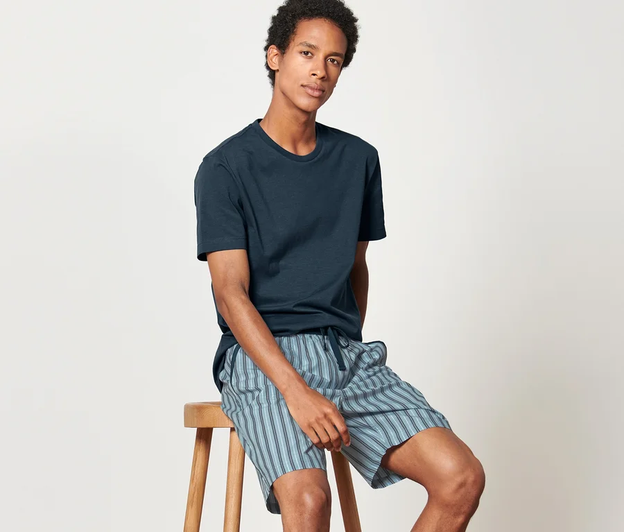 Mann sitzt auf einem Hocker in Shorty-Pyjama mit gewebter Hose und T-Shirt.