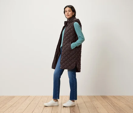 Frau posiert in gesteppter, brauner Weste mit Kapuze, blauem Pullover, Jeans und weißen Schuhen.