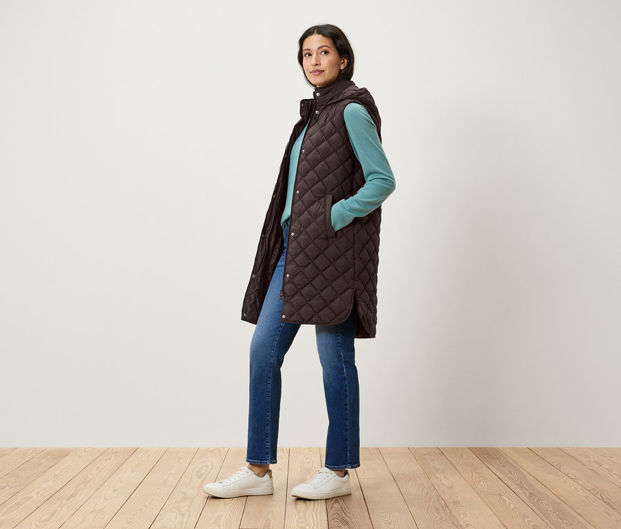 Frau posiert in gesteppter, brauner Weste mit Kapuze, blauem Pullover, Jeans und weißen Schuhen.