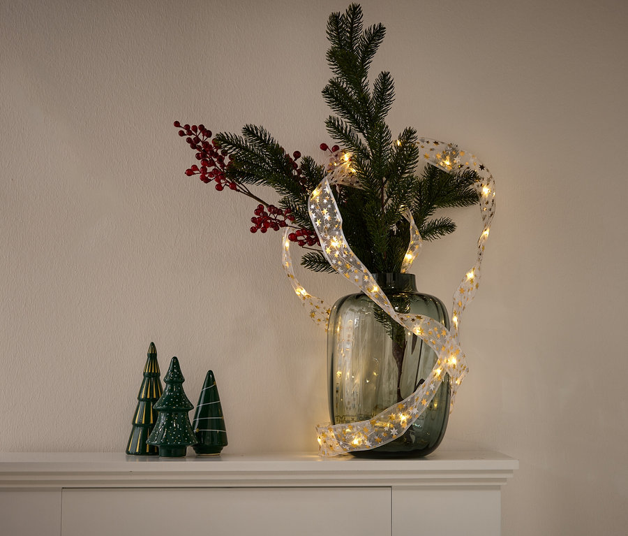 Eine Vase mit Nadelbaumzweigen, roten Beeren und einer Lichterkette mit goldenen Sternen. Links stehen drei grüne Keramikbäumchen.