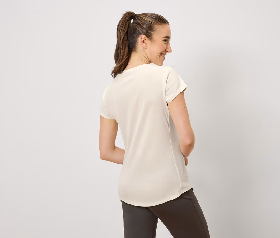 Eine Frau mit braunen, zu einem Pferdeschwanz zusammengebundenen Haaren lächelt und blickt nach links. Sie trägt ein cremefarbenes T-Shirt und braune Hosen.
