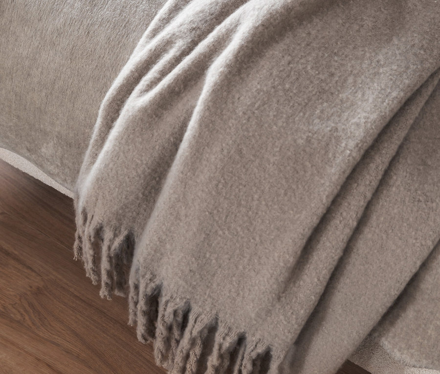 Eine beige Decke mit Fransen liegt auf einem Bett auf einem Holzboden.