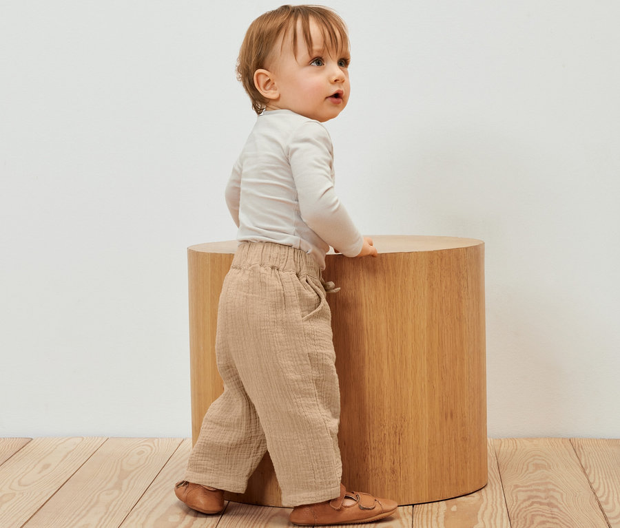 Ein Kind steht neben einem Holzhocker und trägt eine Kinder-Musselin-Hose.
