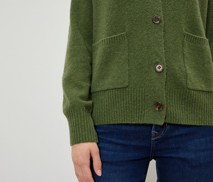 Detailansicht einer Frau, die einen olivgrünen Cardigan mit braunen Knöpfen und Taschen zu dunkelblauen Jeans trägt.