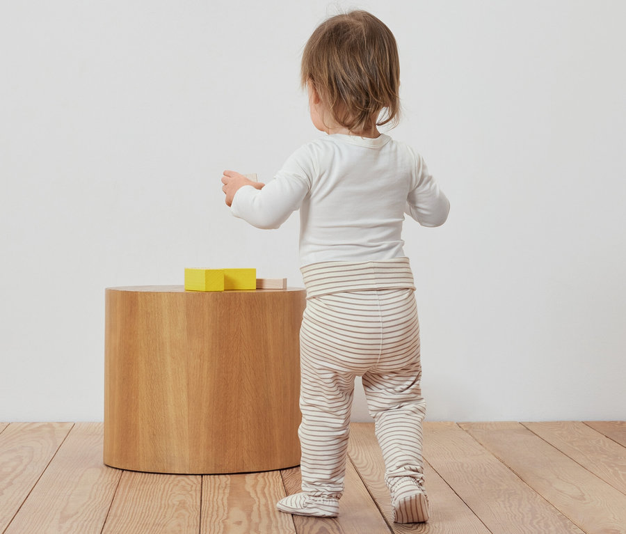 Kleinkind steht mit dem Rücken zur Kamera und spielt mit Spielzeug neben 3 Baby-Strampelhosen.