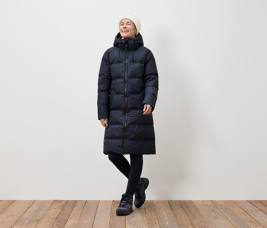 Eine Frau in einem langen, dunkelblauen Daunenmantel, schwarzen Leggings und Winterstiefeln steht auf einem Holzboden und lächelt.