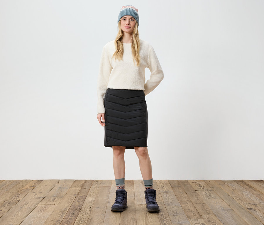 Eine Frau steht auf einem Holzboden und trägt einen cremefarbenen Pullover, einen schwarzen Rock, Stiefel und eine Strickmütze.
