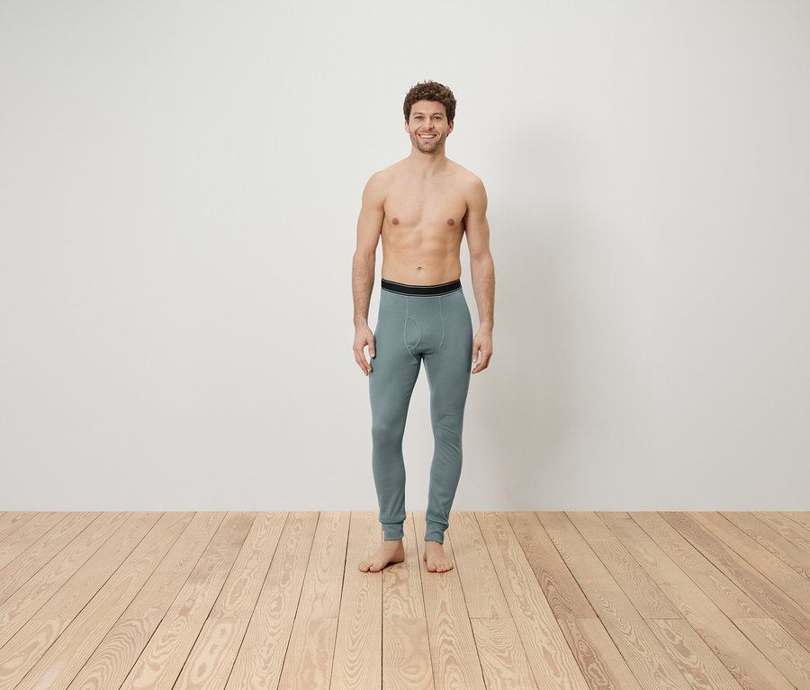 Ein Mann mit freiem Oberkörper steht auf einem Holzboden und trägt graue Leggings.