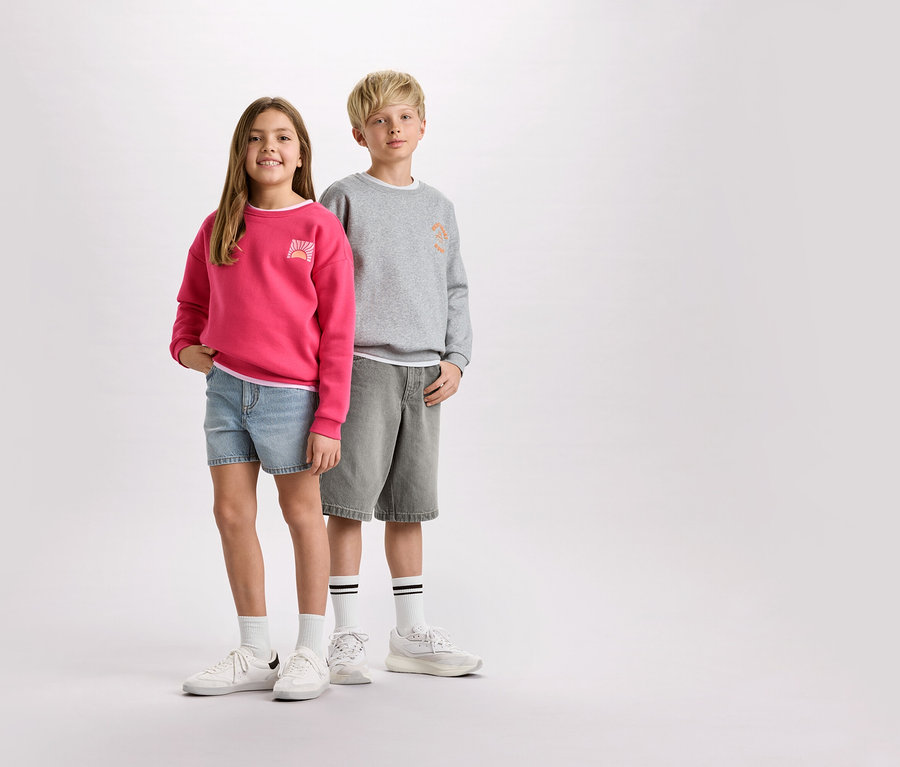 Ein Mädchen mit einem pinken Pullover und Jeansshorts steht neben einem Jungen mit einem grauen Pullover und Shorts.