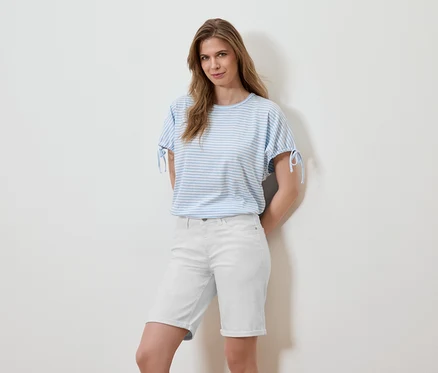 Frau posiert in einem gestreiften, hellblauen T-Shirt und weißen Shorts vor einer weißen Wand.