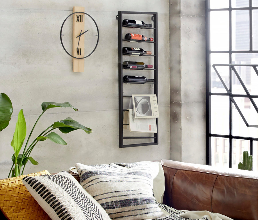 An einer Wand befinden sich eine Uhr, ein Weinregal mit Flaschen und eine Pflanze neben einem Sofa mit Kissen.
