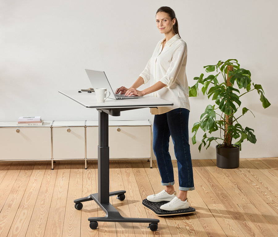 Frau steht an einem höhenverstellbaren Schreibtisch und benutzt einen Laptop, eine Tasse steht daneben.