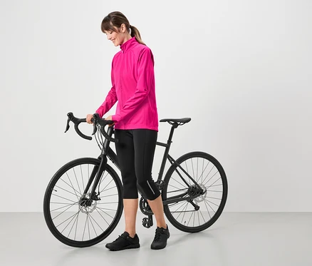 Frau mit pinkfarbener Jacke, schwarzen Radlerhosen und schwarzen Schuhen, die neben einem schwarzen Fahrrad steht.