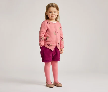 Ein kleines Mädchen mit blonden Haaren trägt eine rosa Strickjacke mit Kirschmuster, violette Shorts, rosa Strumpfhosen und Ballerinas.