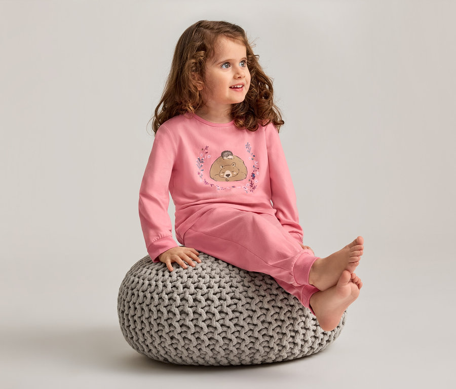 Mädchen mit lockigem Haar sitzt barfuß auf einem grauen Strickhocker und trägt einen rosa Schlafanzug mit Bärenmotiv.