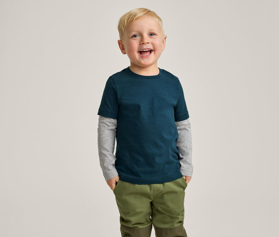 Ein lächelnder kleiner Junge mit blondem Haar trägt ein dunkelblaugrünes T-Shirt über einem grauen Langarmshirt und olivgrüne Hosen, die Hände in den Taschen.
