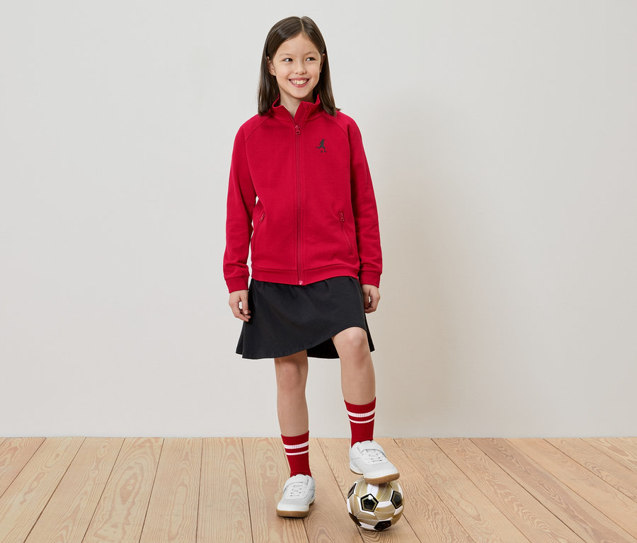 Mädchen steht mit einem Fuß auf einem Fußball, trägt eine rote Jacke, einen schwarzen Rock, weiße Schuhe und rot-weiße Socken.
