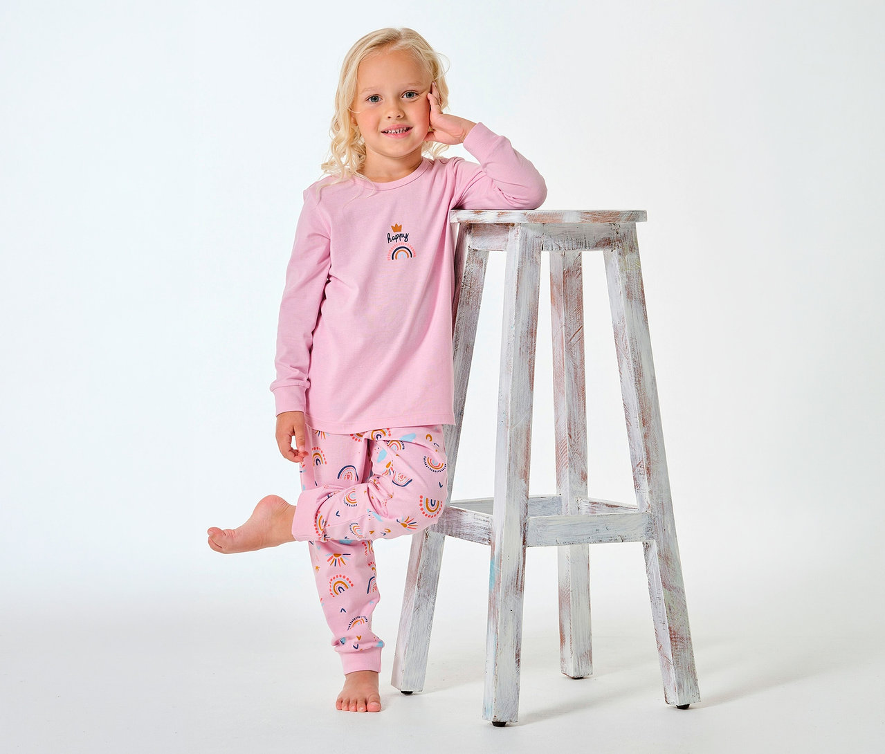 Mädchen im langen SCHIESSER Kinder-Pyjama mit Regenbogen-Motiv, an einem Hocker lehnend.