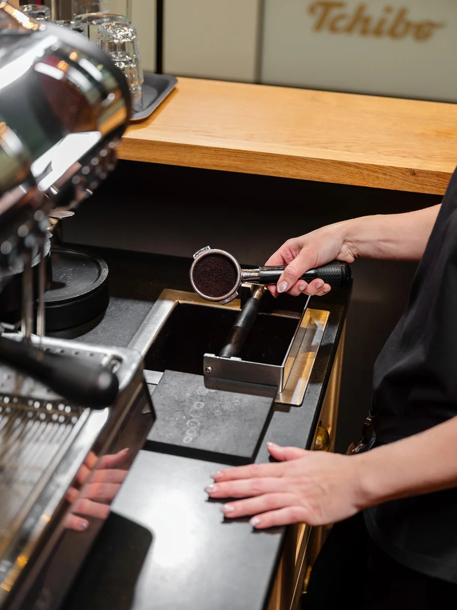 Eine Person hält einen Siebträger mit Kaffeepulver über einen Ausklopfkasten neben einer Kaffeemaschine.