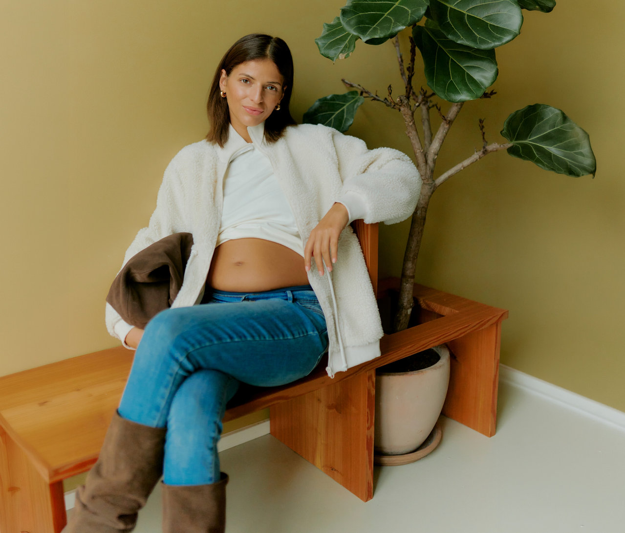 Schwangere sitzt auf Bank neben Pflanze und trägt MAMALICIOUS 3-in-1-Teddyjacke »MLCARLY« offen über weißem Shirt und Jeans.