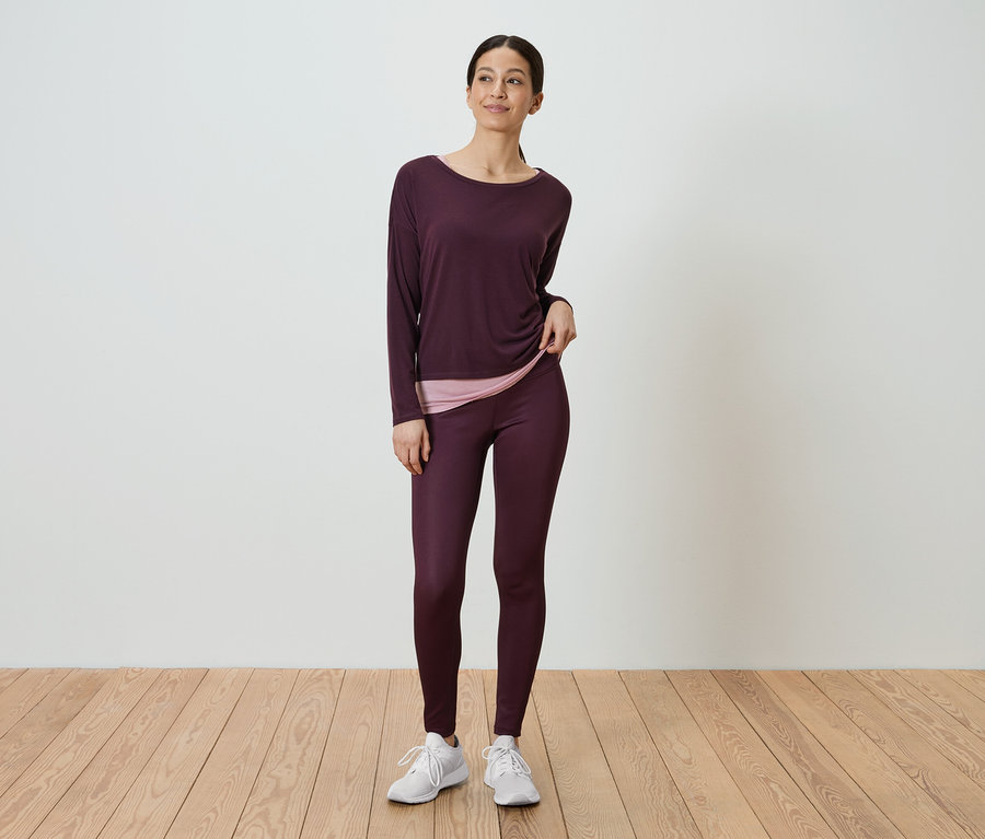 Frau steht und trägt ein langärmliges, auberginefarbenes Hemd und auberginefarbene Leggings.