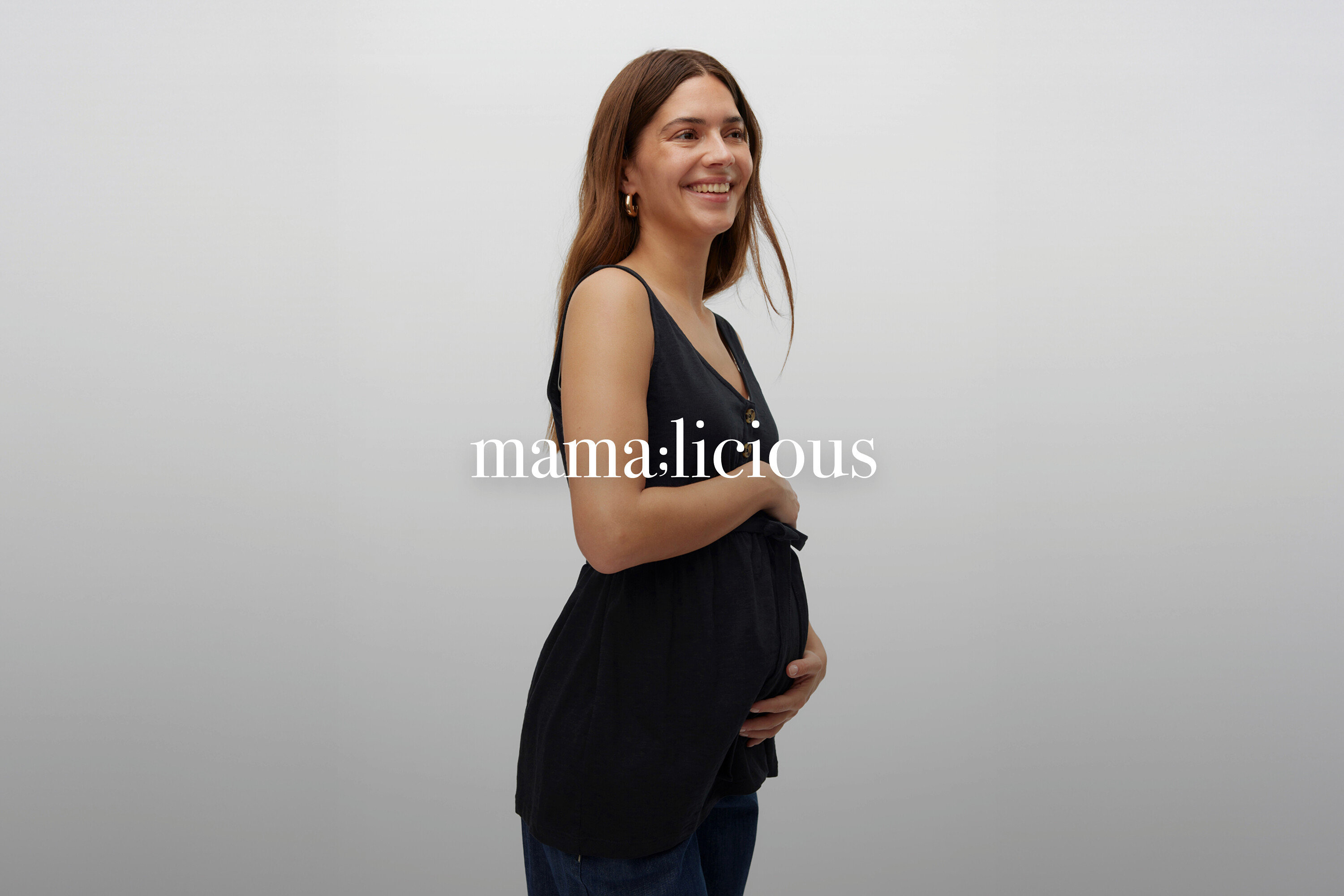 Lächelnde Schwangere mit langen braunen Haaren, die ein ärmelloses, geknöpftes, schwarzes Umstandsoberteil und Jeans trägt.