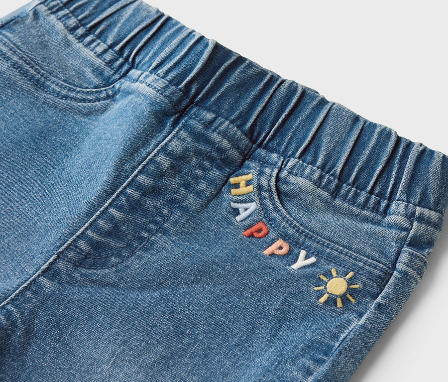 Nahaufnahme von Jeans mit Gummibund und dem Wort "HAPPY" und einem Sonnen-Emoji auf der Tasche.