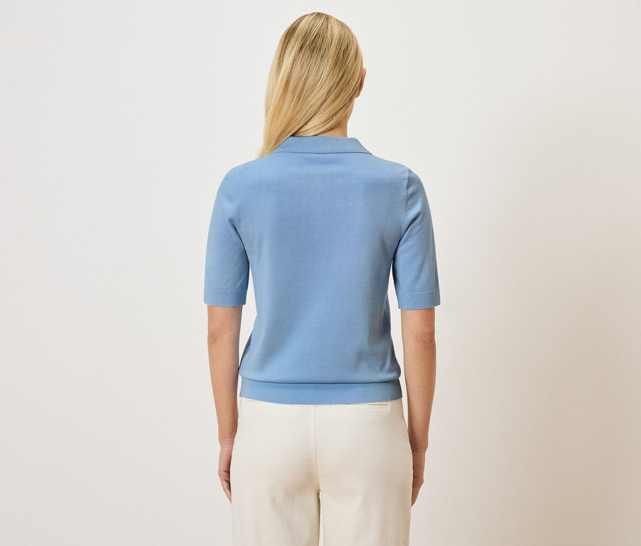 Rückansicht einer Frau mit blonden Haaren, die ein hellblaues Poloshirt und eine helle Hose trägt.