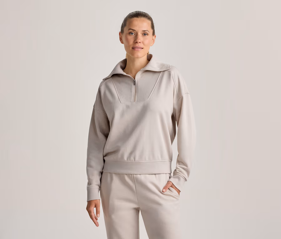 Frau steht, trägt ein hellbeiges Half-Zip-Sweatshirt und passende Jogginghose, eine Hand in der Tasche, blickt nach vorne.