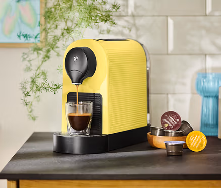 Eine gelbe Kaffeemaschine füllt eine Tasse mit Kaffee, Kapseln liegen daneben.