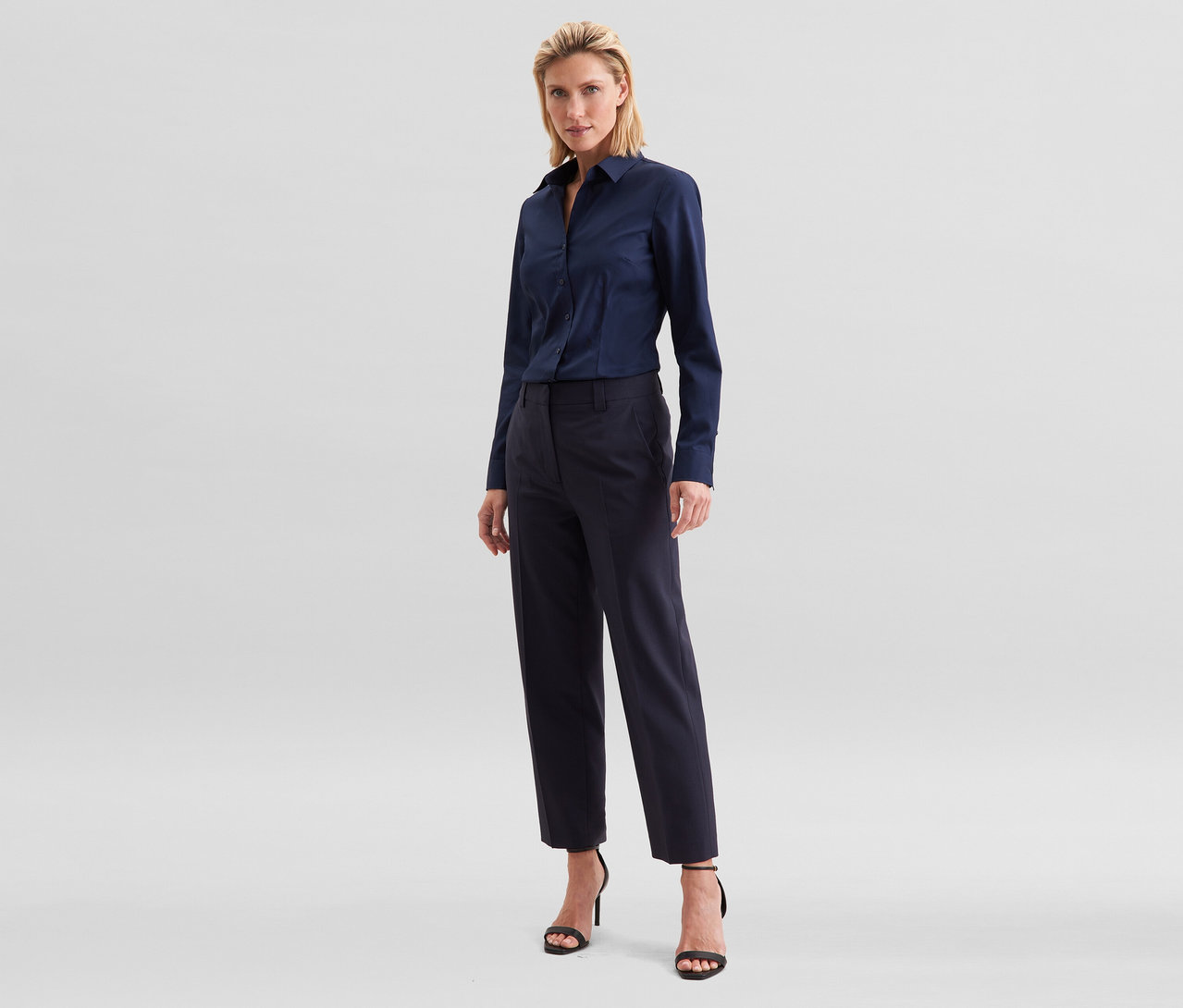 Blonde Frau steht in einem dunkelblauen Langarmhemd, einer dunkelblauen Cropped-Hose und schwarzen Sandaletten mit hohen Absätzen.