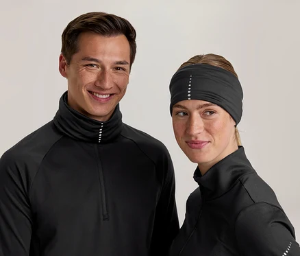 Ein lächelnder Mann und eine Frau tragen schwarze Sportoberteile mit hohem Kragen und reflektierenden Punkten. Die Frau trägt auch ein schwarzes Stirnband mit reflektierenden Punkten.
