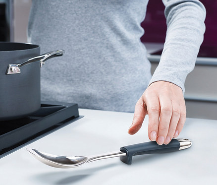 Hand greift nach einem Joseph Joseph Elevate Steel Schöpflöffel mit dunkelgrauem Griff, dessen Kopf erhöht aufliegt, neben einem dunkelgrauen Topf auf einer weißen Arbeitsplatte.