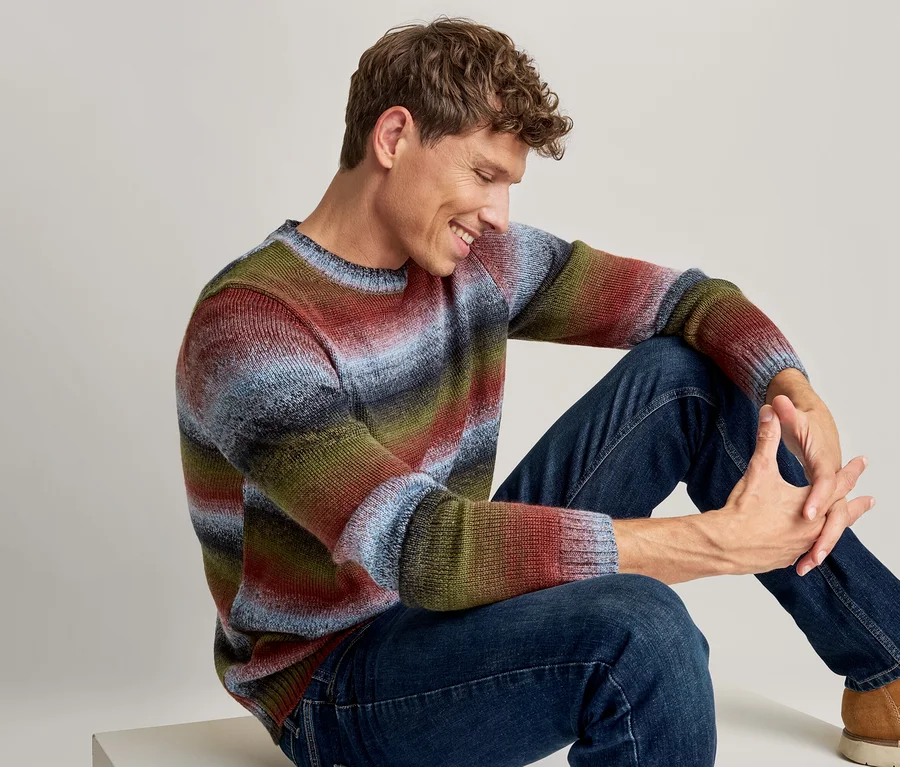 Sitzender Mann, der lächelt und nach unten blickt, trägt einen mehrfarbigen gestreiften Pullover, dunkelblaue Jeans und hellbraune Stiefel.