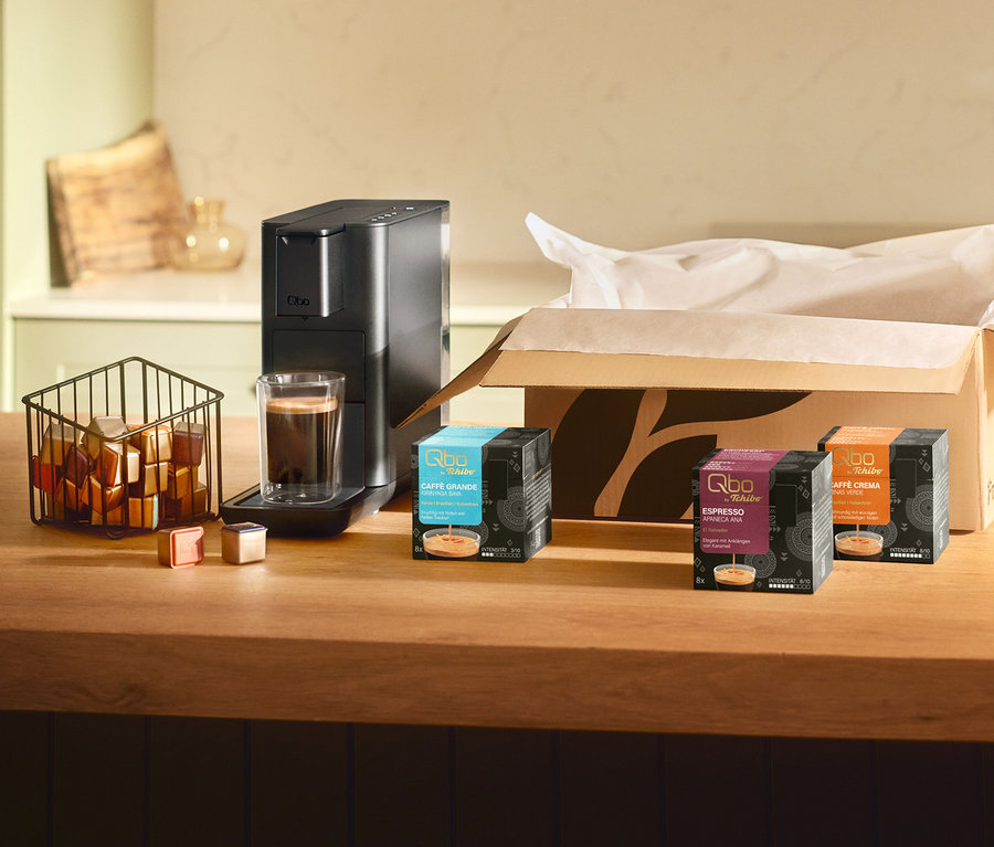 Schwarze Qbo Iconic Dark Chrome Kaffeemaschine mit Kaffee in einem Glas, daneben ein Korb mit Kapseln und drei Qbo Kaffeekapselpackungen: Qbo Caffè Grande KIRINYAGA BAYA, Qbo Espresso APANECA ANA und Qbo Caffè MINAS VERDE.