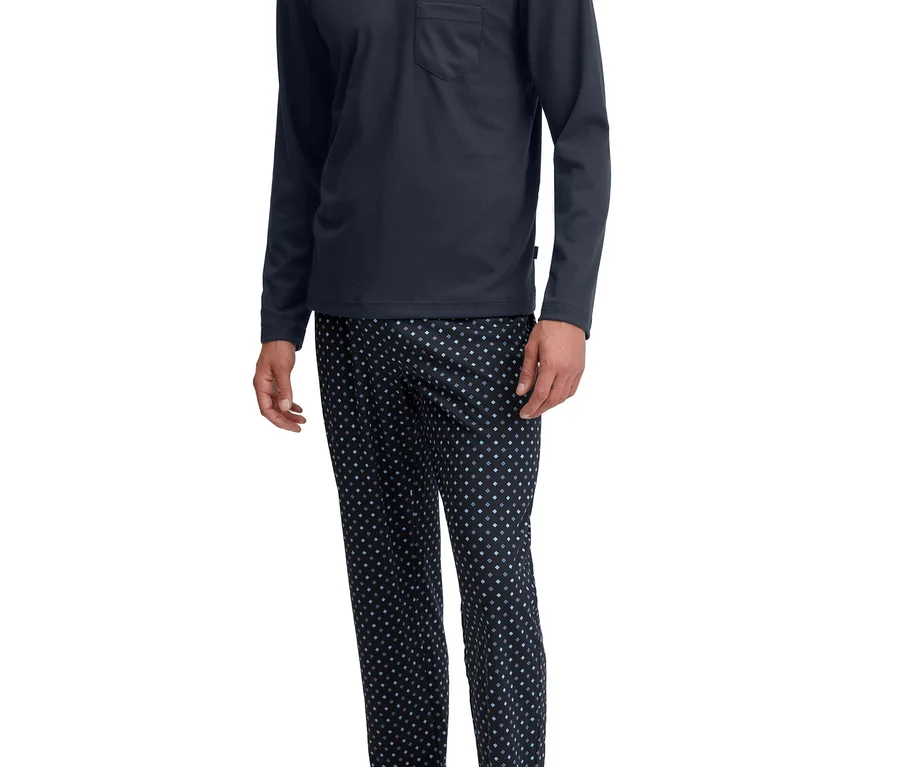 Mann steht lächelnd in einem dunkelblauen Langarm-V-Ausschnitt-Pyjamaoberteil mit Brusttasche und dunkel gemusterter Pyjamahose, barfuß.