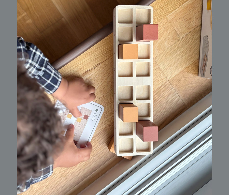 Kinderhände halten eine Musterkarte neben einem small foot Montessori-Bundle 2 Holzspielzeug mit orangefarbenen und braunen Blöcken auf einem Holzboden.