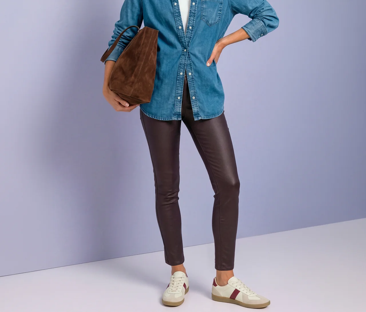 Eine Person trägt eine blaue Jeansbluse, eine Street One Hose in Lederoptik in Rot und cremefarbene Sneaker mit roten Streifen, eine braune Wildledertasche haltend.