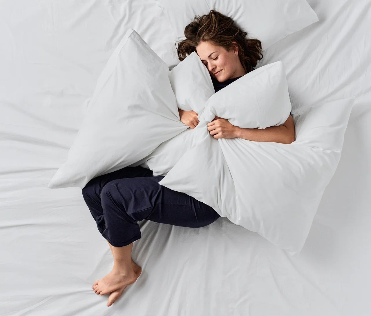 Frau schläft auf einem weißen Bett, umarmt ein großes weißes Kissen. Sie trägt eine dunkelblaue CALIDA »Deepsleepwear Balancing« Hose.