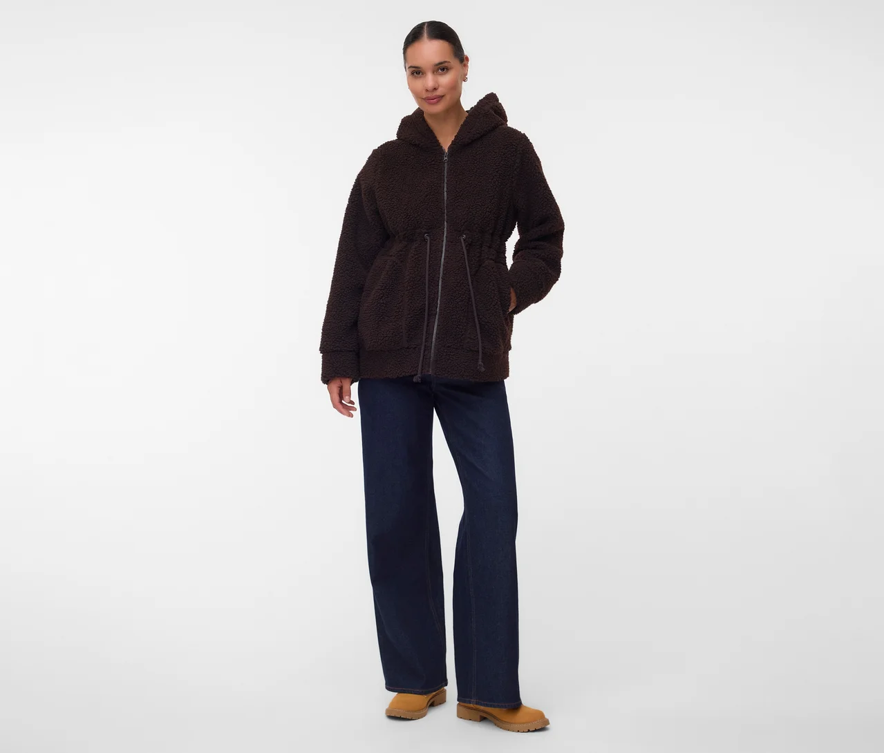 Frau in einer braunen Kapuzen-Fleecejacke mit Kordelzug in der Taille, dunkelblauen weiten Jeans und beigen Stiefeln, die mit einer Hand in der Tasche steht.