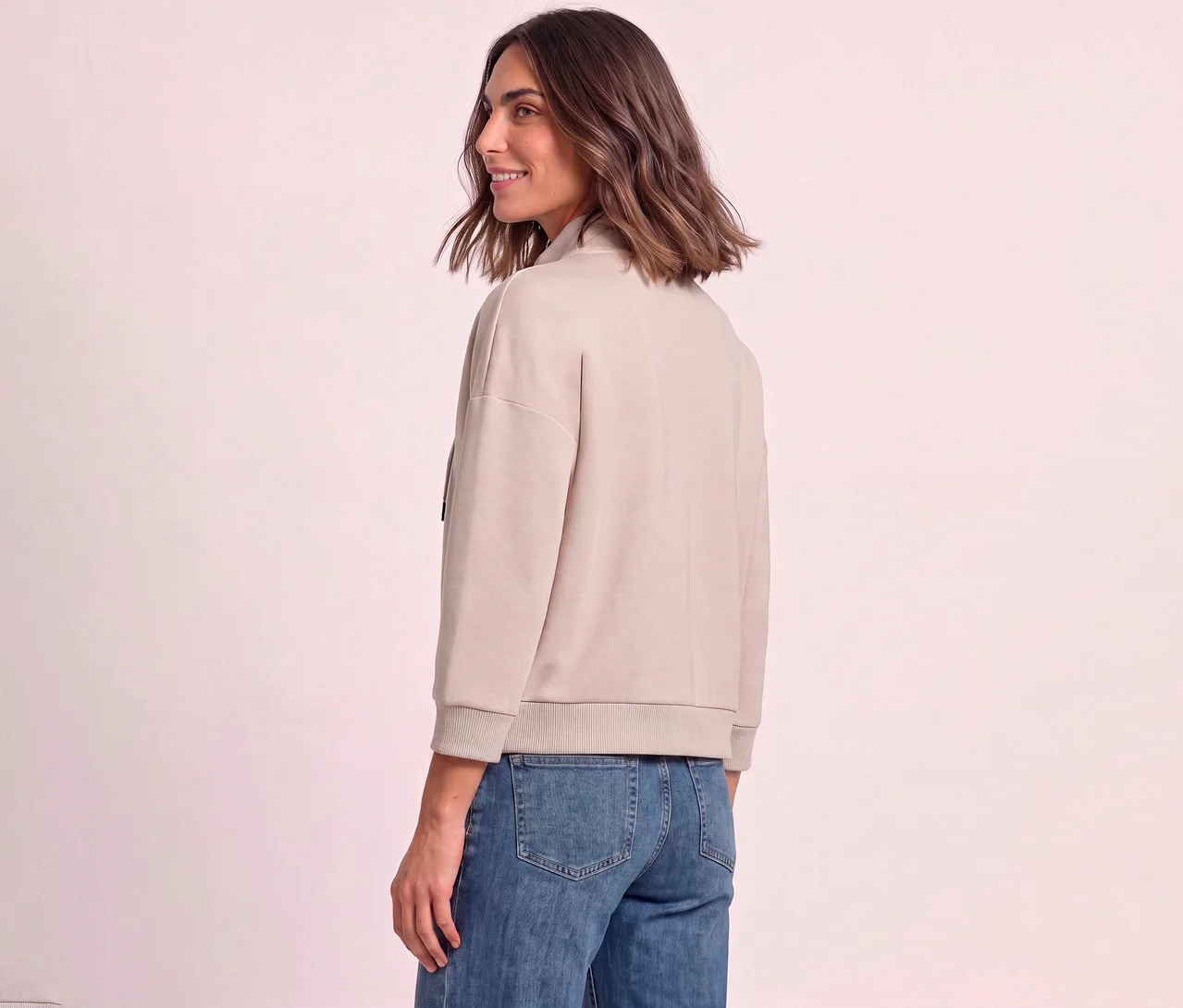 Frau trägt eine hellbeige Street One Sweatjacke und blaue Jeans, blickt über ihre Schulter und lächelt.
