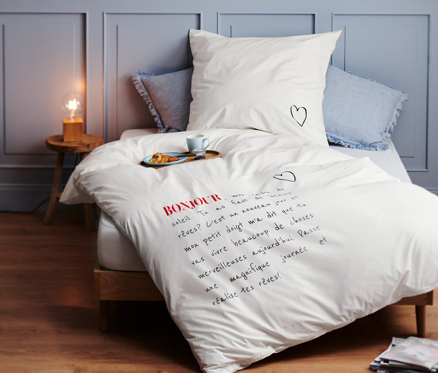 Weiße Premium-Baumwoll-Bettwäsche in Normalgröße mit Bonjour-Schriftzug und kleinem Herz auf dem Kissen liegt auf einem Bett.