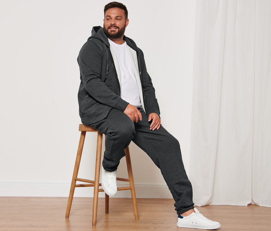 Ein Mann sitzt auf einem Holzhocker und trägt eine anthrazit melierte Kapuzenjacke, 2 weiße T-Shirts mit Rundhalsausschnitt, eine Sweathose und Sneaker.