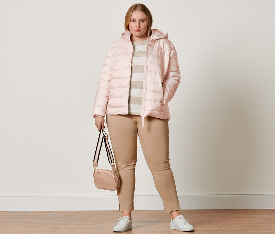 Frau trägt eine rosa Steppjacke mit Kapuze, eine Stretchhose und Ledersneaker. Sie hat eine Umhängetasche und ein gestreiftes T-Shirt.