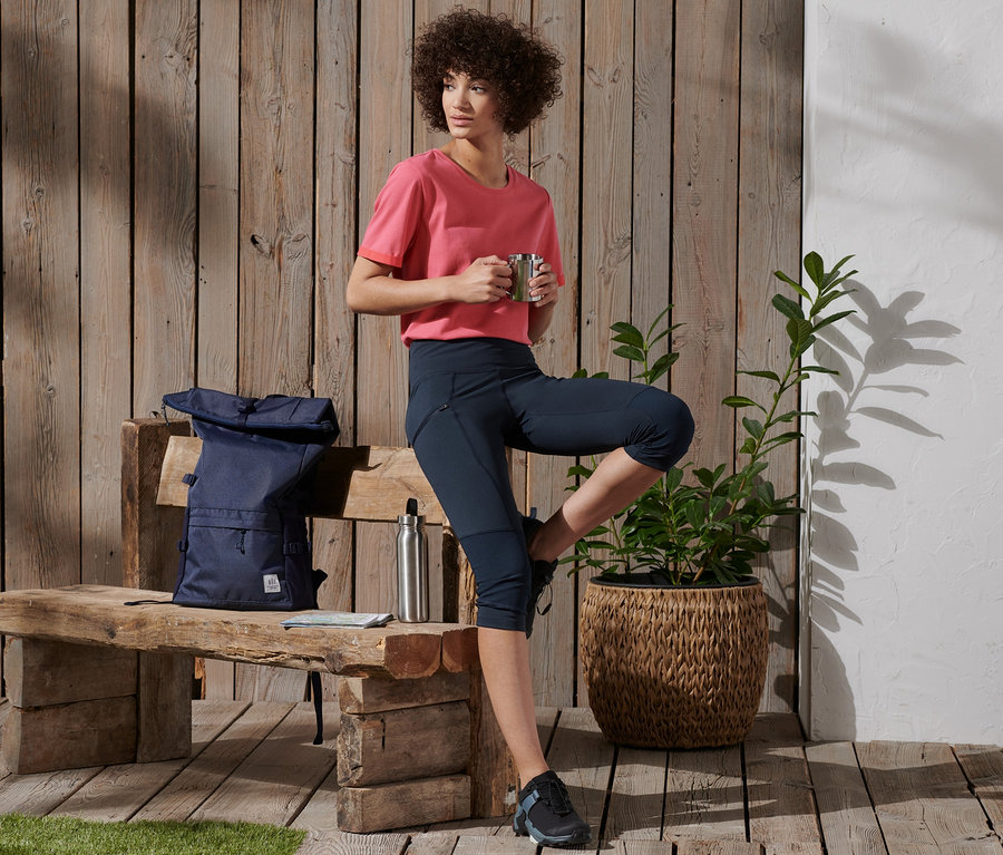 Eine Frau hält eine Tasse und steht mit einem Bein auf einer Bank. Auf der Bank liegen ein Rucksack und eine Wasserflasche. Sie trägt eine 3/4-Trekking-Tights.